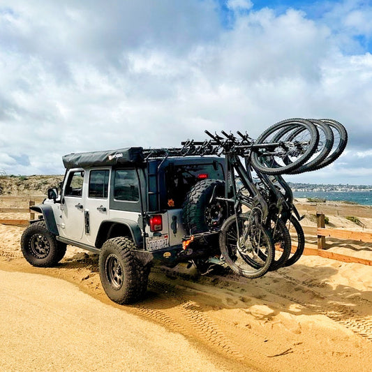 A jeep with a vertical bike rack mounted on the roof, parked on a sandy surface with a body of water and sky in the background.